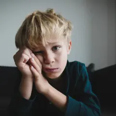 Chłopiec z blond włosami, zamyślony, z rękami przy twarzy. Jego wyraz twarzy może sugerować niektóre objawy aspergera, jak trudności w wyrażaniu emocji.