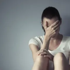 Kobieta w białej koszulce siedzi przy szarej ścianie, zakrywając twarz dłońmi. Czasem każdy potrzebuje chwili miansec, by zebrać myśli.