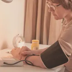Kobieta z polsartem mierzy ciśnienie. Na stole leżą tabletki i budzik.