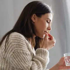 Kobieta w swetrze bierze tabletkę i trzyma szklankę wody.