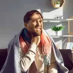 Mężczyzna z zarostem, okryty kocem i szalikiem, odczuwa ostry ból gardła przy przełykaniu, choć nie ma gorączki.
