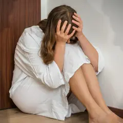 Kobieta w białej koszuli siedzi skulona przy drzwiach, trzymając się za głowę.