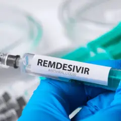 Ręka w niebieskiej rękawiczce trzyma strzykawkę z płynem **remdesivir**. W tle widać inne strzykawki.