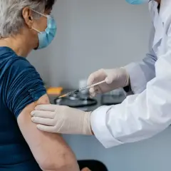 Starsza pani otrzymuje szczepionkę na covid. Lekarz w białym fartuchu i rękawiczkach podaje zastrzyk.