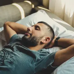 Mężczyzna leży na łóżku z rękami za głową, oczy zamknięte, w stonowanych barwach.