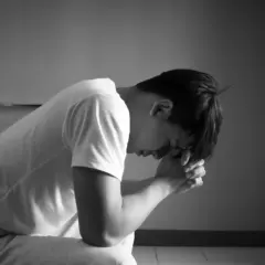 Mężczyzna siedzi na łóżku, pogrążony w melancholii, z rękami złożonymi przed twarzą.