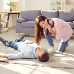 Kobieta wzywa pomoc przez telefon, klęcząc obok nieprzytomnego mężczyzny leżącego na dywanie.