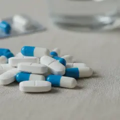 Kapsułki i tabletki w kolorze białym i niebieskim, część opakowania leków, szklanka wody w tle. NLPZ.