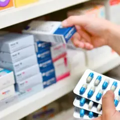 Ręka wybiera z półki aptecznej pudełko leku, a w drugiej dłoni trzyma blistry z kapsułkami, być może zawierającymi cefuroksym.