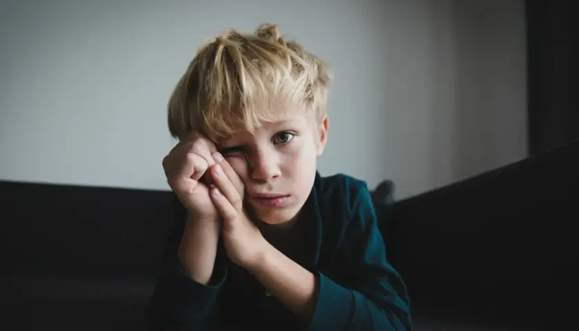 Chłopiec z blond włosami, zamyślony, z rękami przy twarzy. Jego wyraz twarzy może sugerować niektóre objawy aspergera, jak trudności w wyrażaniu emocji.
