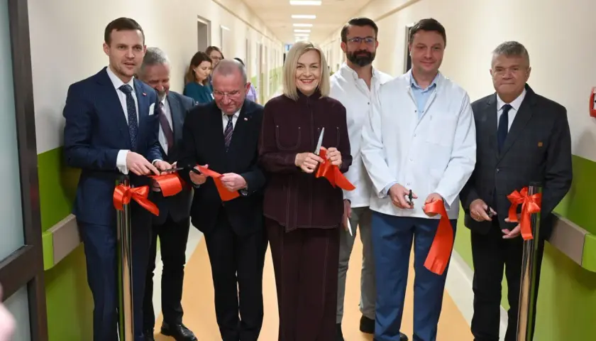 Grupa osób w garniturach i kitlach przecina czerwone wstęgi w szpitalnym korytarzu.