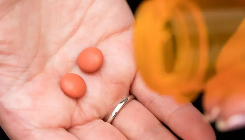 Dwie pomarańczowe tabletki, być może Inflanor Max, leżą na dłoni obok otwartego pojemnika.