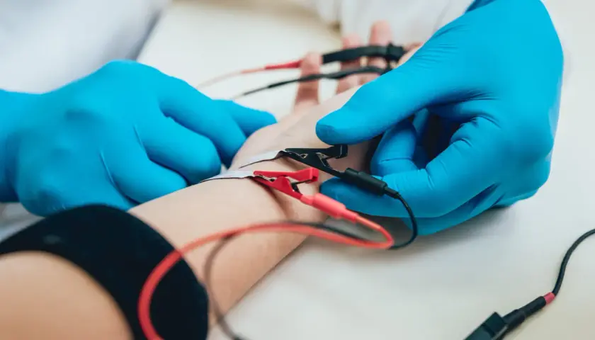 Lekarz w niebieskich rękawiczkach podłącza elektrody do ręki pacjenta podczas badania EMG.