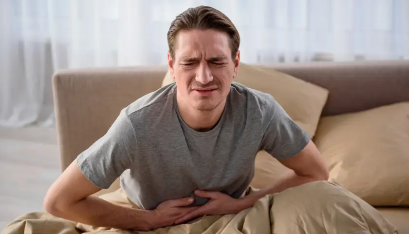 Mężczyzna w łóżku trzymający się za brzuch z wyrazem bólu na twarzy.
