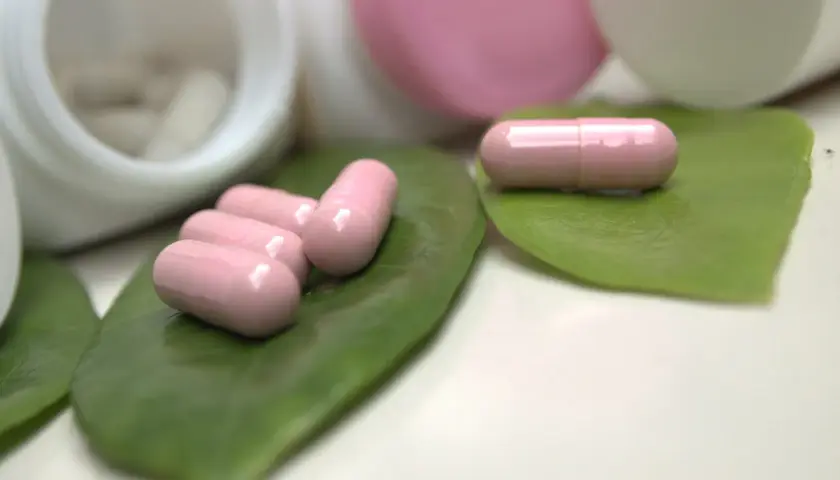 Różowe kapsułki, być może zawierające ketoprofen, leżą na zielonych liściach obok otwartych pojemników z lekami.