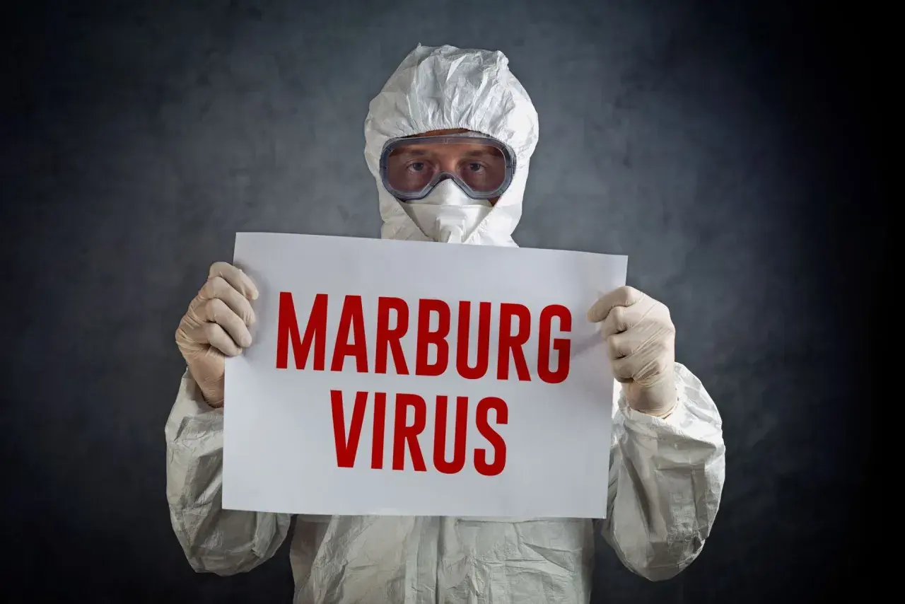 Osoba w kombinezonie ochronnym trzyma tabliczkę z napisem "MARBURG VIRUS".