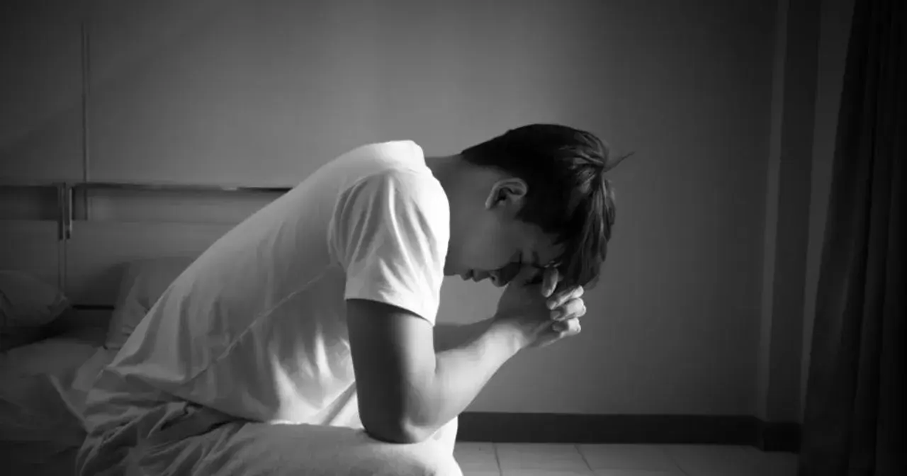 Mężczyzna siedzi na łóżku, pogrążony w melancholii, z rękami złożonymi przed twarzą.
