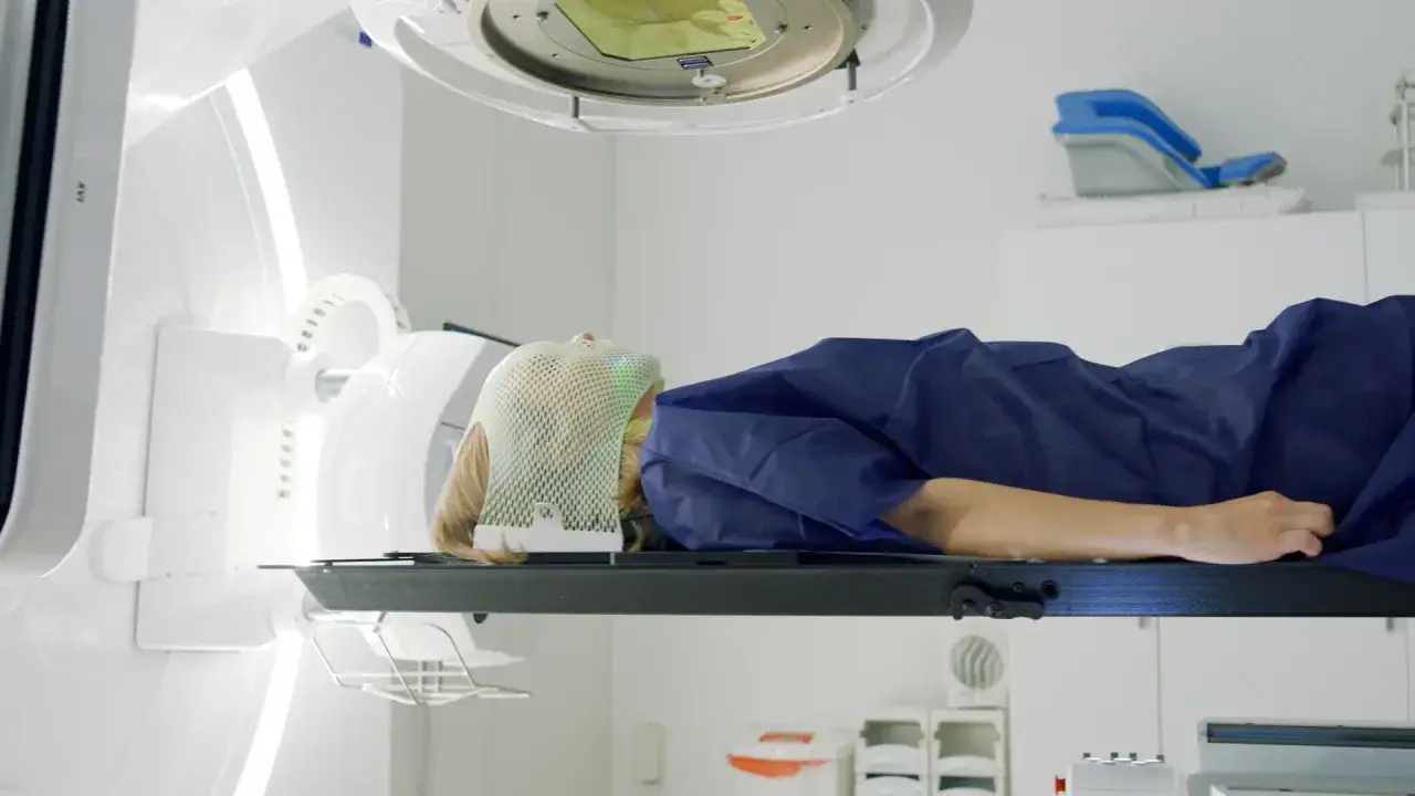 Pacjentka leży na stole zabiegowym, ma na twarzy maskę pozycjonującą. Trwa radioterapia, precyzyjne leczenie.