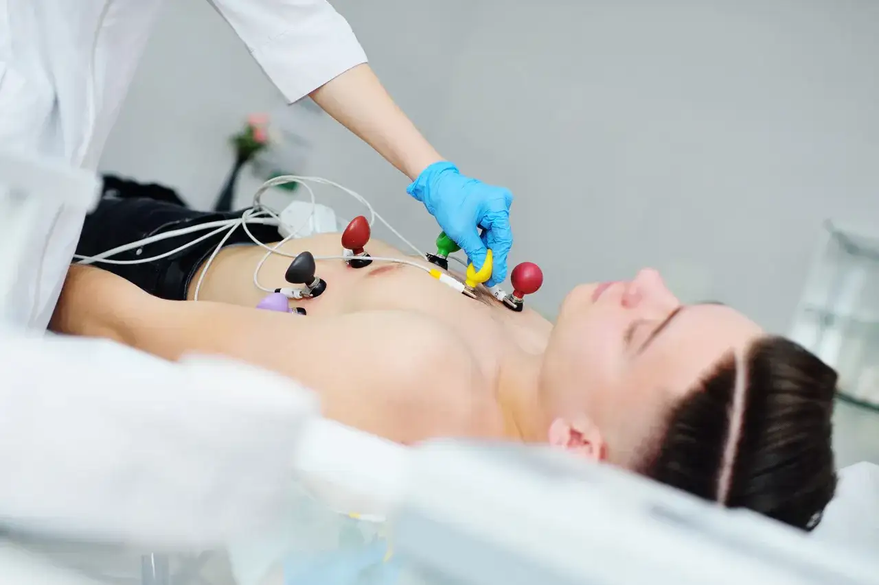 Pacjent leży z elektrodami EKG na klatce piersiowej. Lekarz w niebieskiej rękawiczce podłącza przewody, monitorując pracę serca, co może być pomocne przy ocenie wpływu leków takich jak amiodaron.